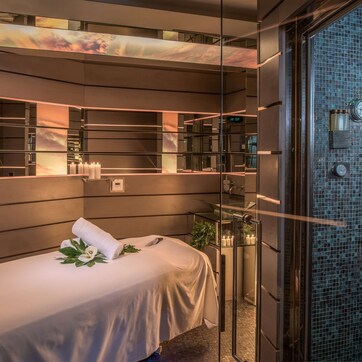 A modern spa room with a massage table, a large mirror, and a glass shower enclosure.