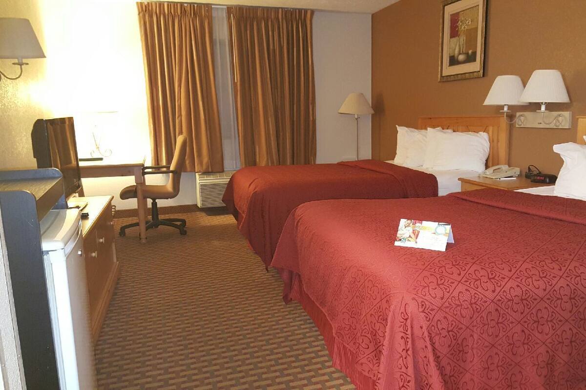 Quarto de hotel com duas camas, uma escrivaninha com cadeira, telefone e luminária.