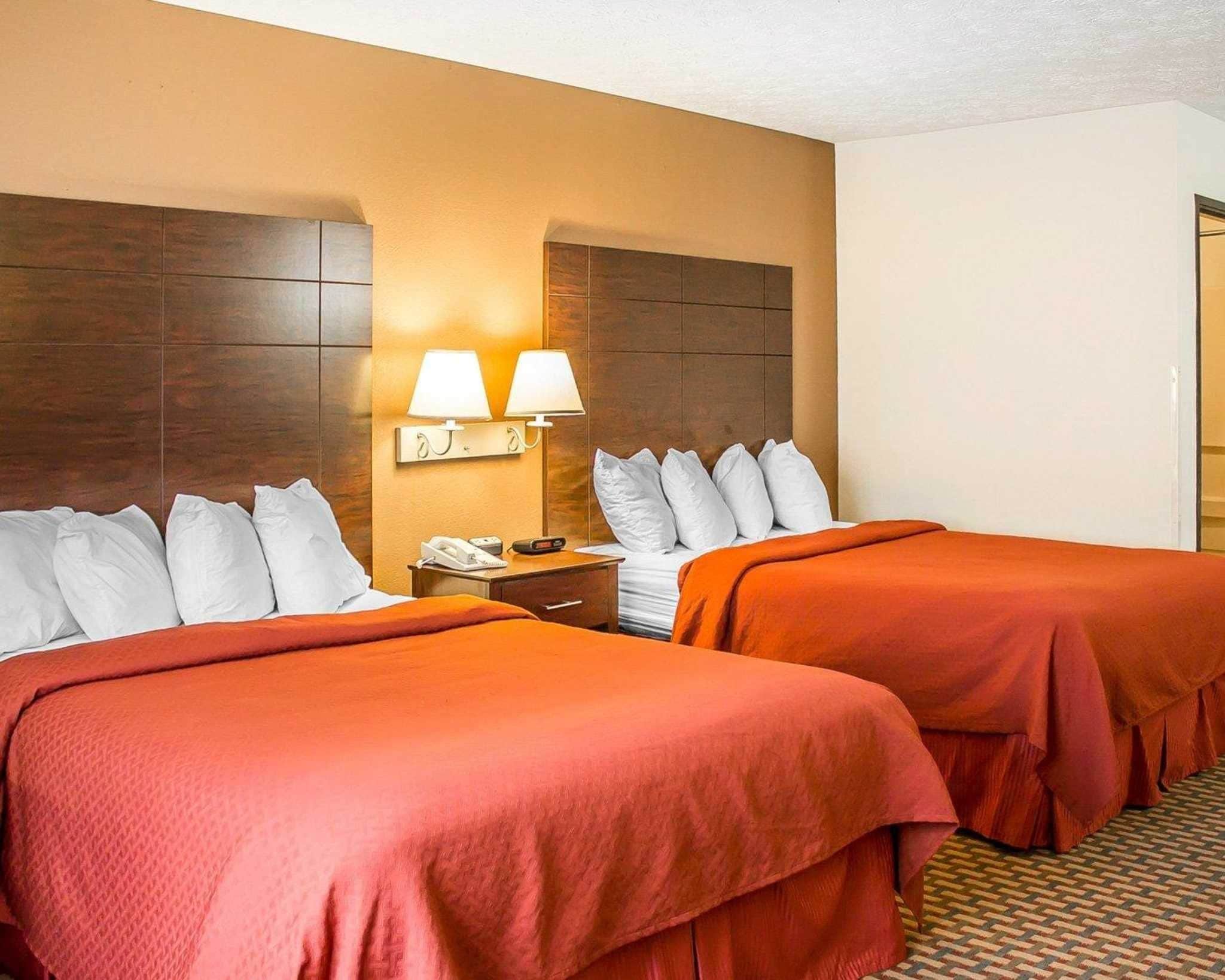 A hotel room with two beds, a nightstand with a phone and lamp, and a wall-mounted light fixture.