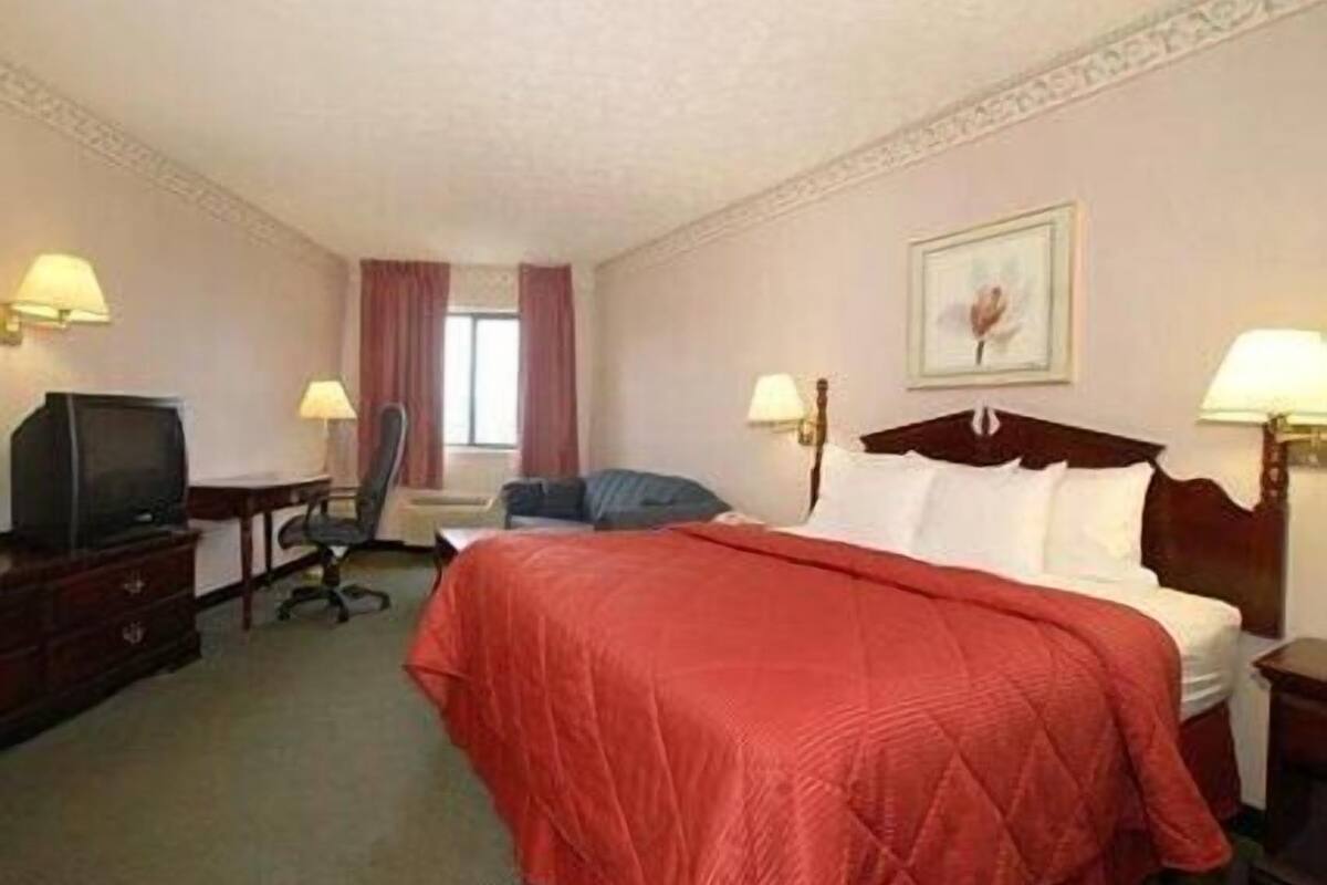 Quarto de hotel com uma cama grande, televisão, escrivaninha com luminária e uma cadeira.