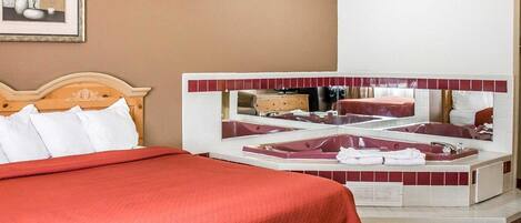 Habitación de hotel con una cama grande, una zona de estar con un sofá rojo, una bañera y un espejo que refleja la habitación.