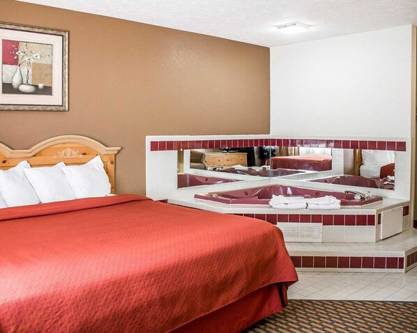 A hotel room with a large bed, a sitting area with a red sofa, a bathtub, and a mirror reflecting the room.