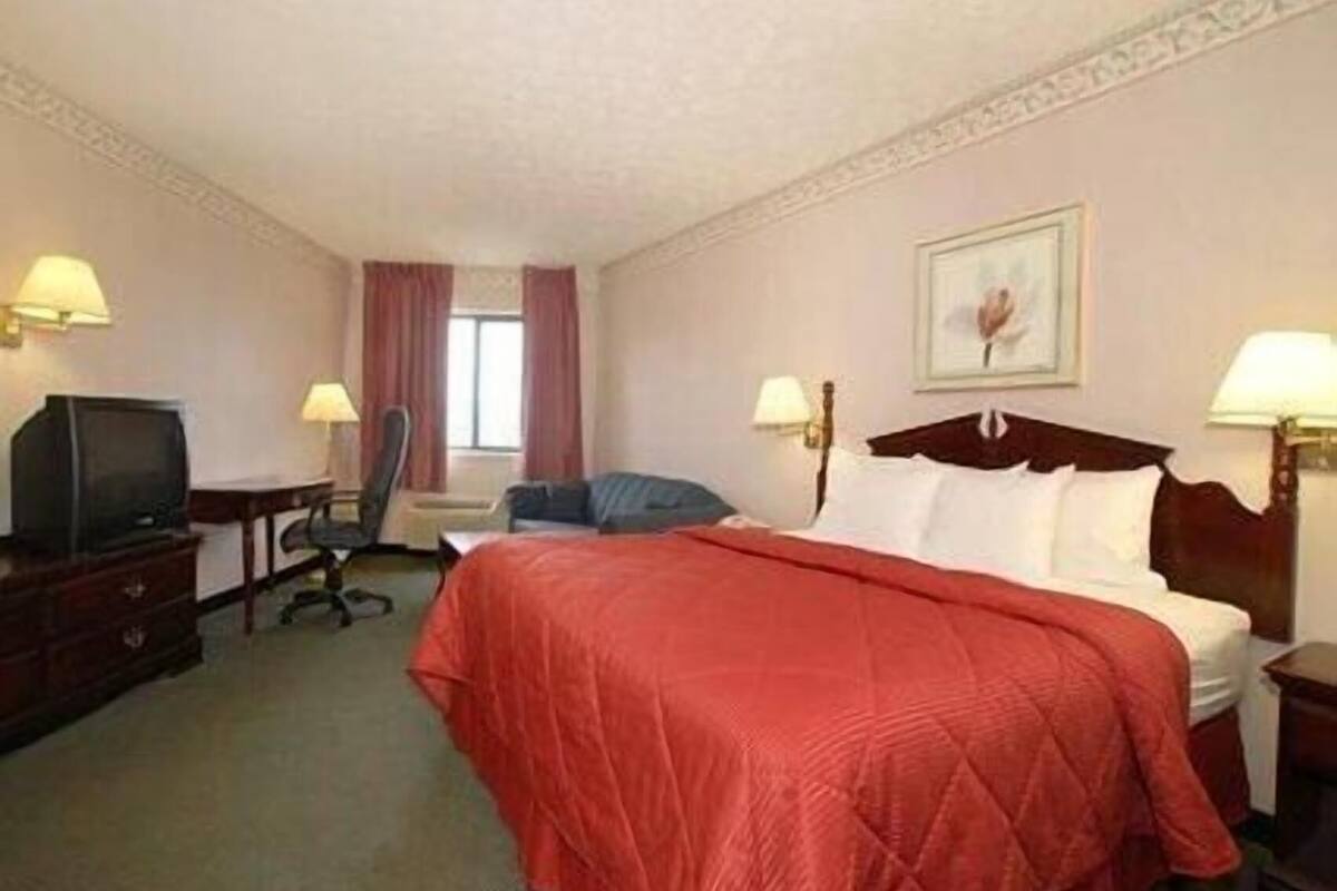 Quarto de hotel com cama, secretária com televisão, uma cadeira e duas luminárias.