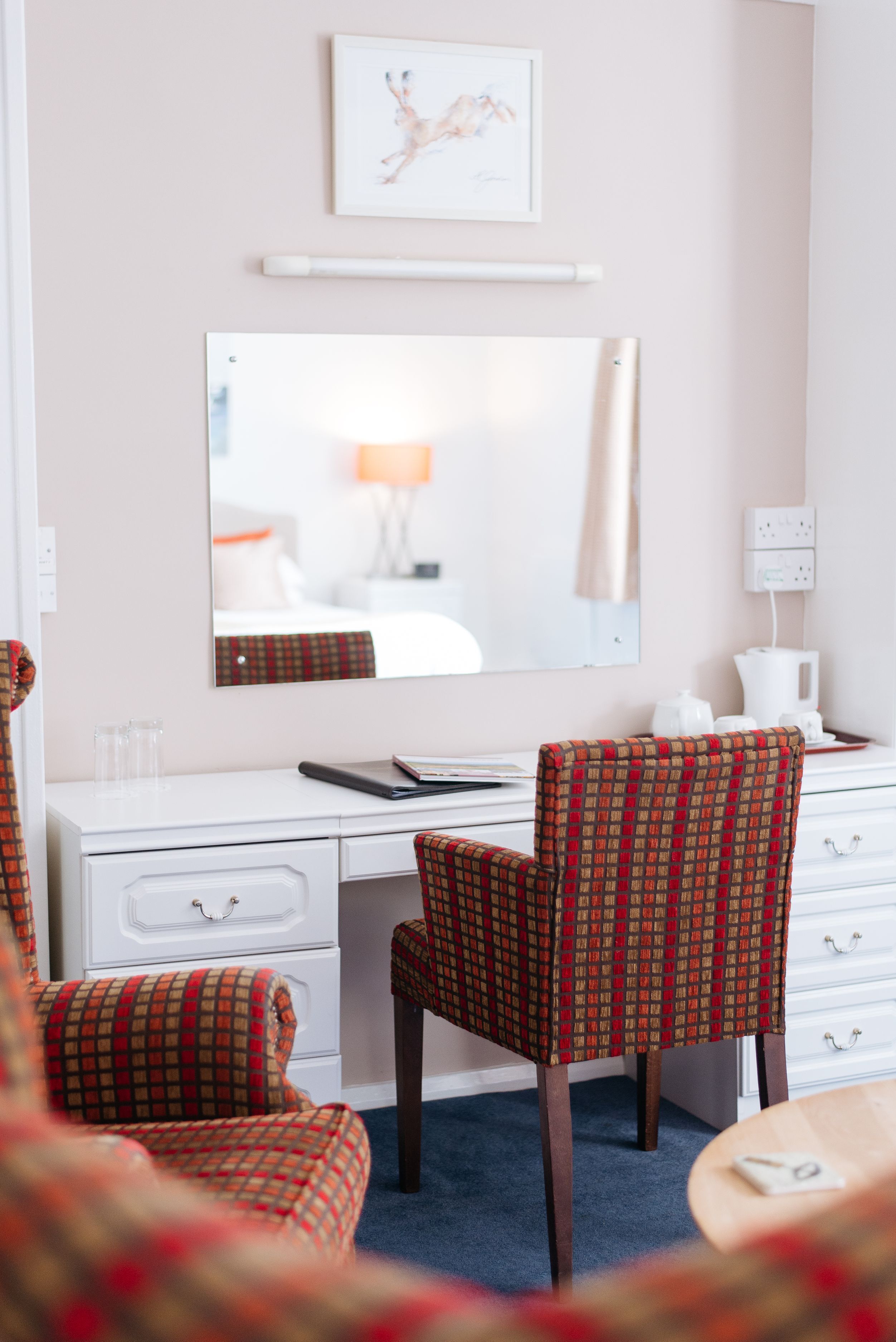 A hotel room with a white desk, a chair with a plaid pattern, a mirror, a framed picture on the wall, and a blue carpet.