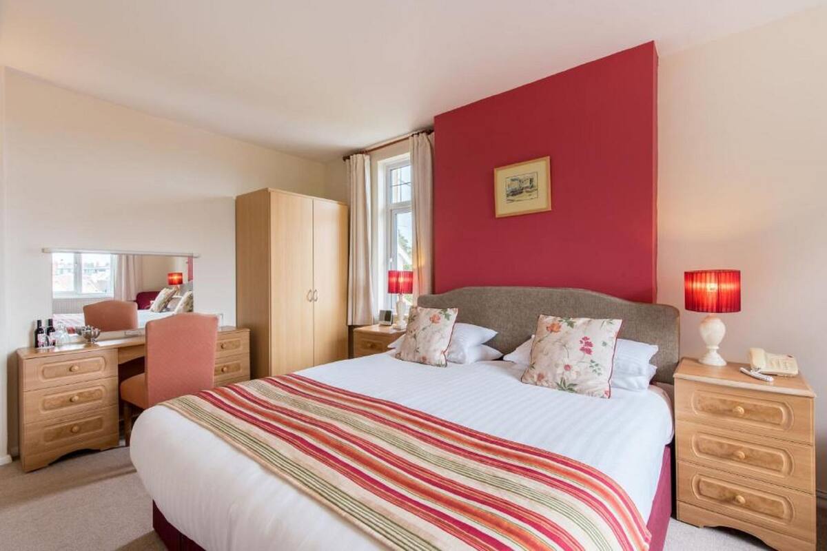 A hotel room with a large bed, a desk, a wardrobe, and a nightstand with a lamp.