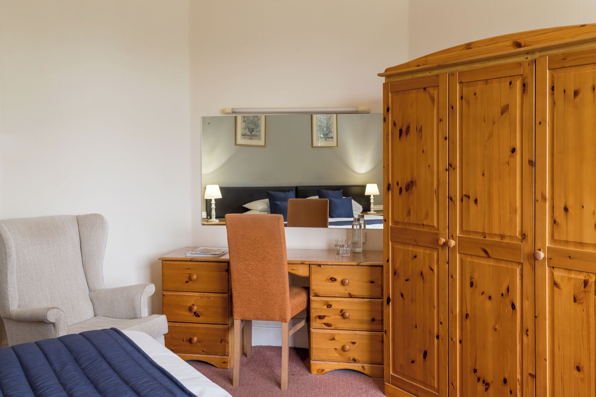A hotel room with a bed, a desk with a chair, a wardrobe, and a mirror.