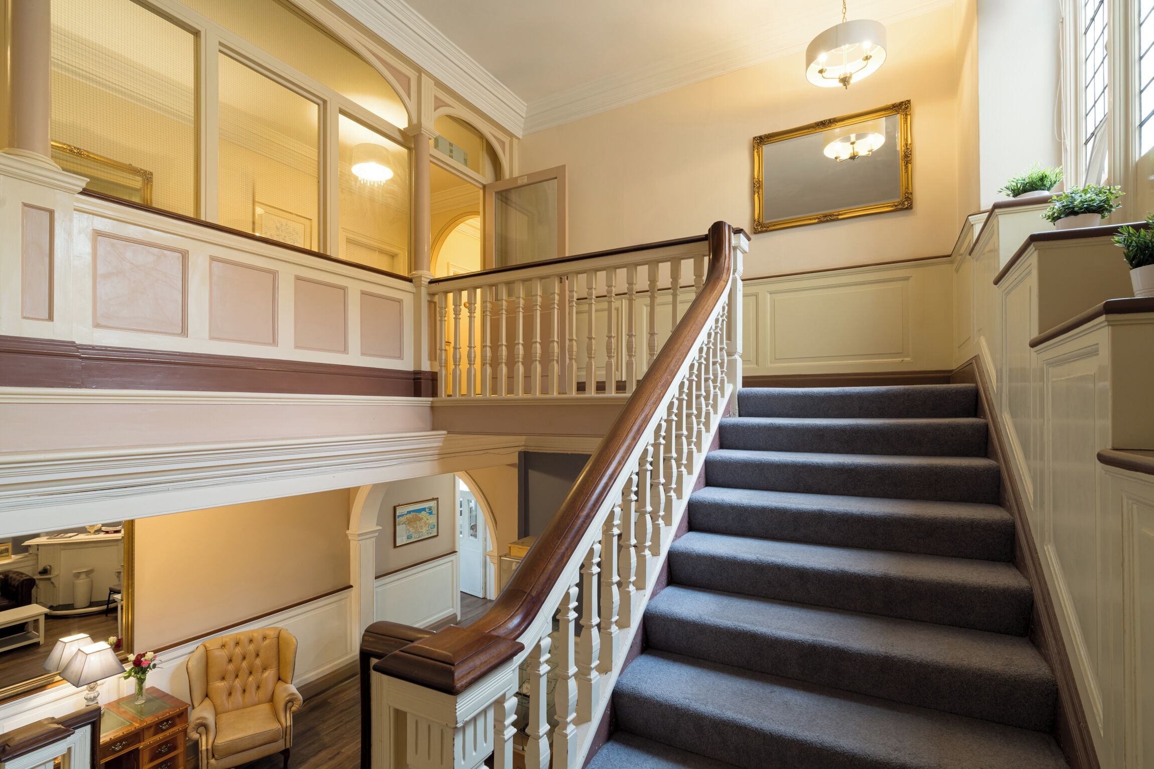 A grand staircase with a wooden handrail and a chandelier, leading to a second-floor landing with a view of the interior.