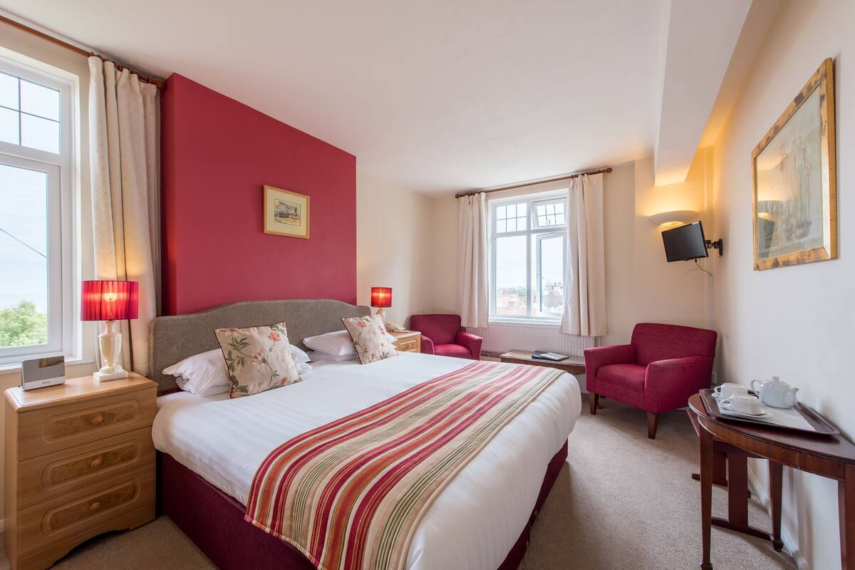 A hotel room with a large bed, a desk, a chair, a nightstand, a lamp, a television, and a painting on the wall.