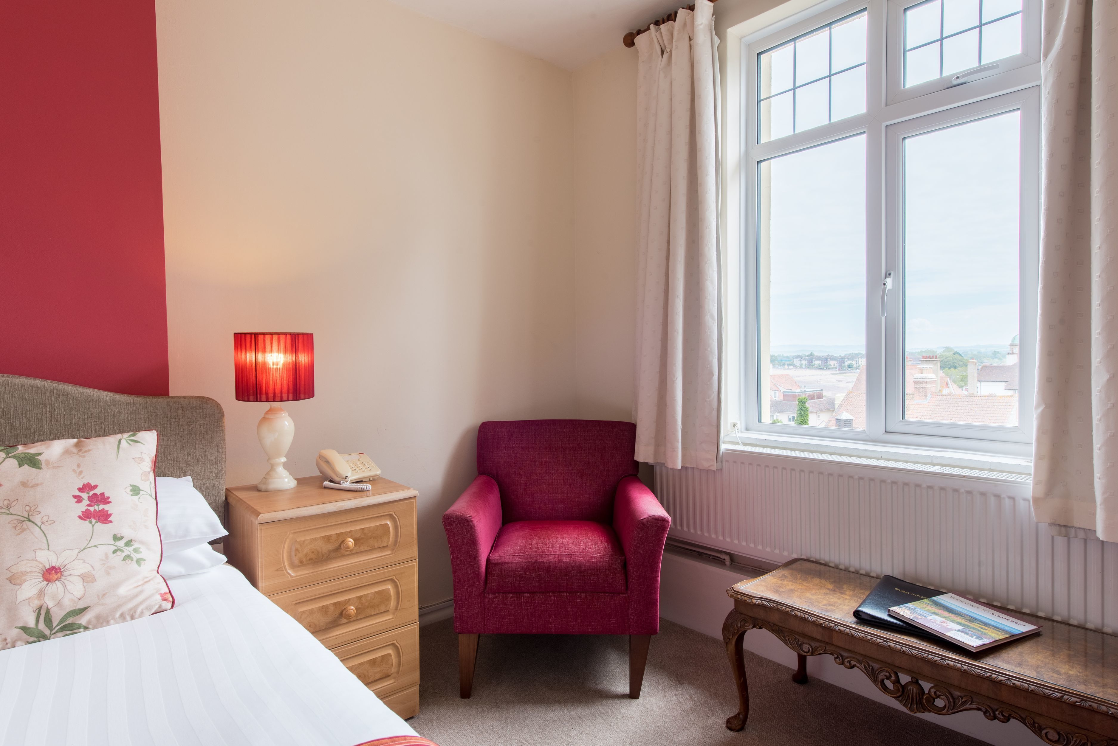 Une chambre à coucher avec un lit, une table de chevet avec une lampe, une chaise, une petite table avec un livre et une fenêtre donnant sur un beau paysage.