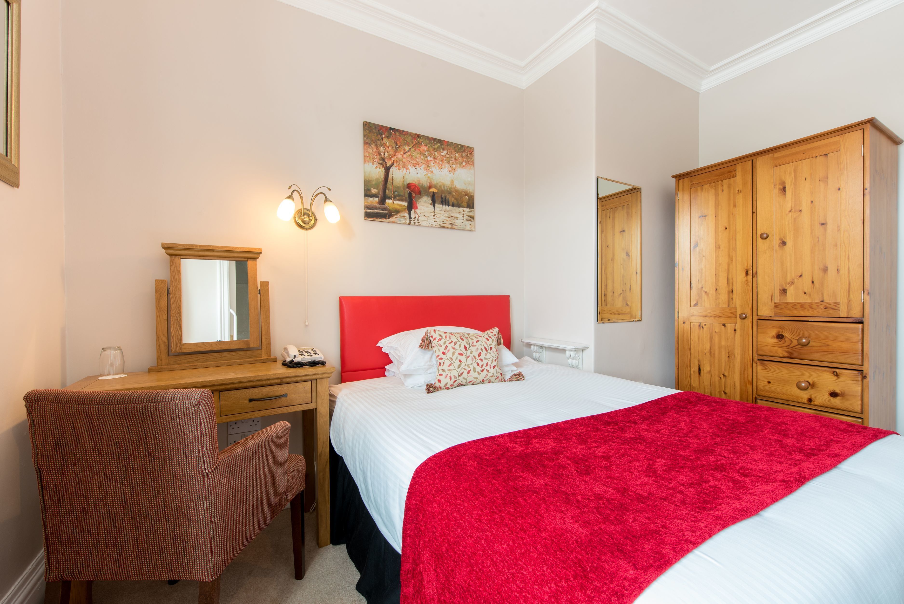 A bedroom with a bed, a desk, a chair, a wardrobe, a painting, and a mirror.