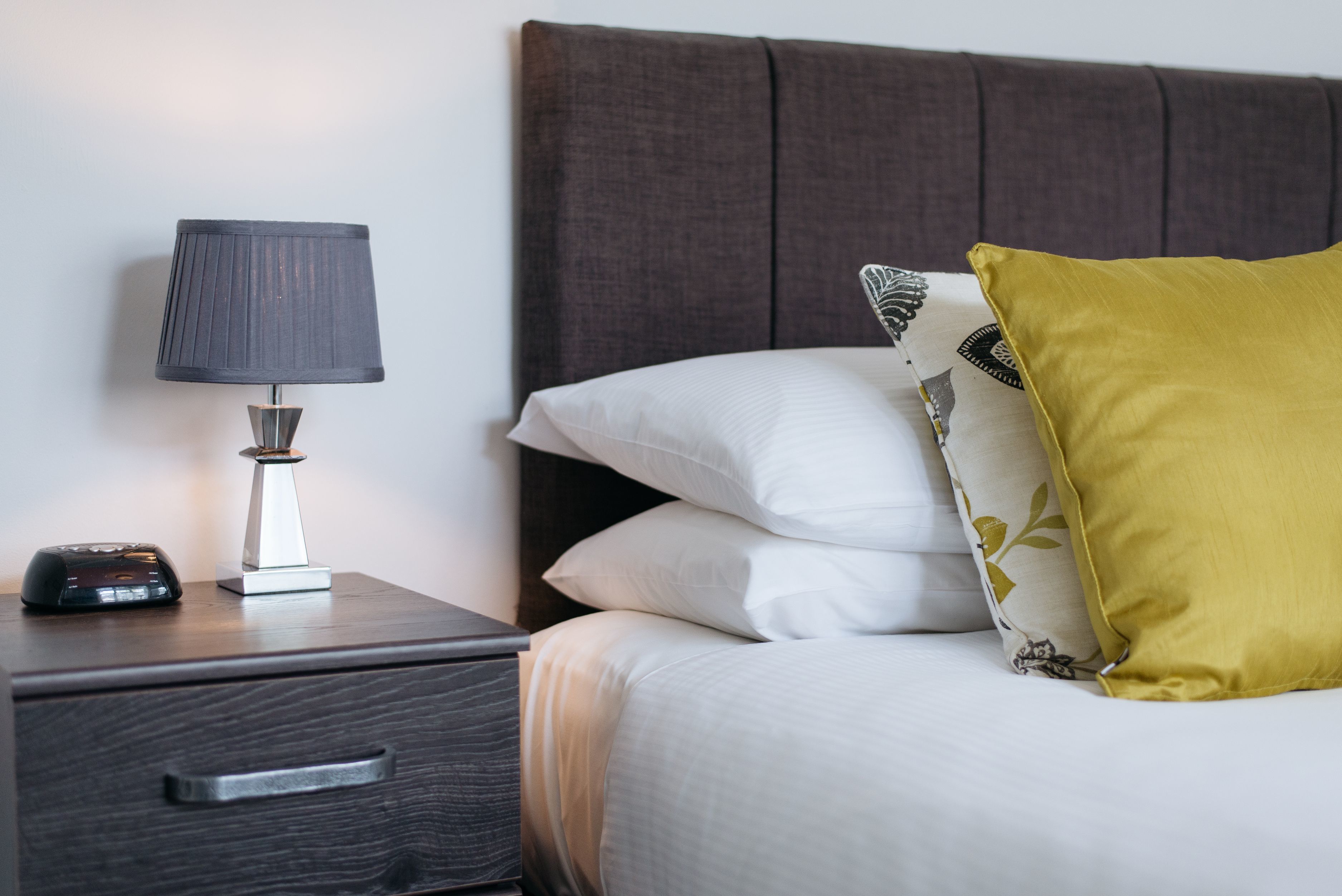 A neatly made bed with white and patterned pillows, a bedside lamp, and a dark wooden nightstand with a drawer.