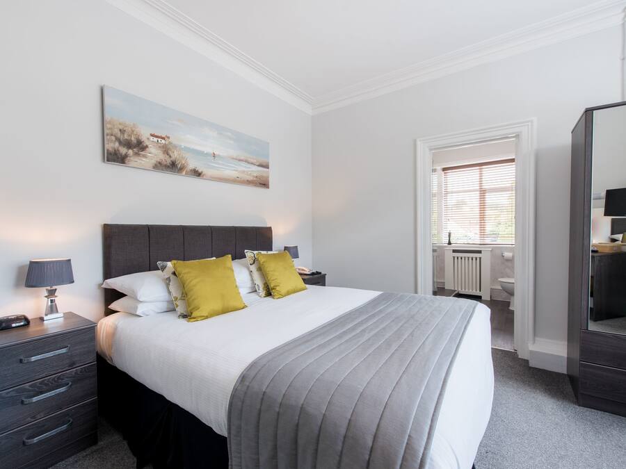 A neatly arranged bedroom with a bed, bedside tables, a nightstand, a painting on the wall, and a bathroom visible through an open door.