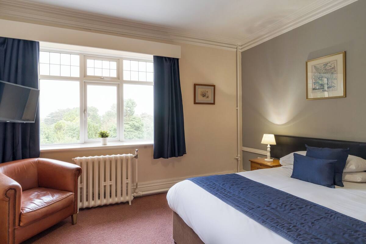 A bedroom with a bed, a chair, a radiator, a window with curtains, a nightstand with a lamp, and a framed picture on the wall.