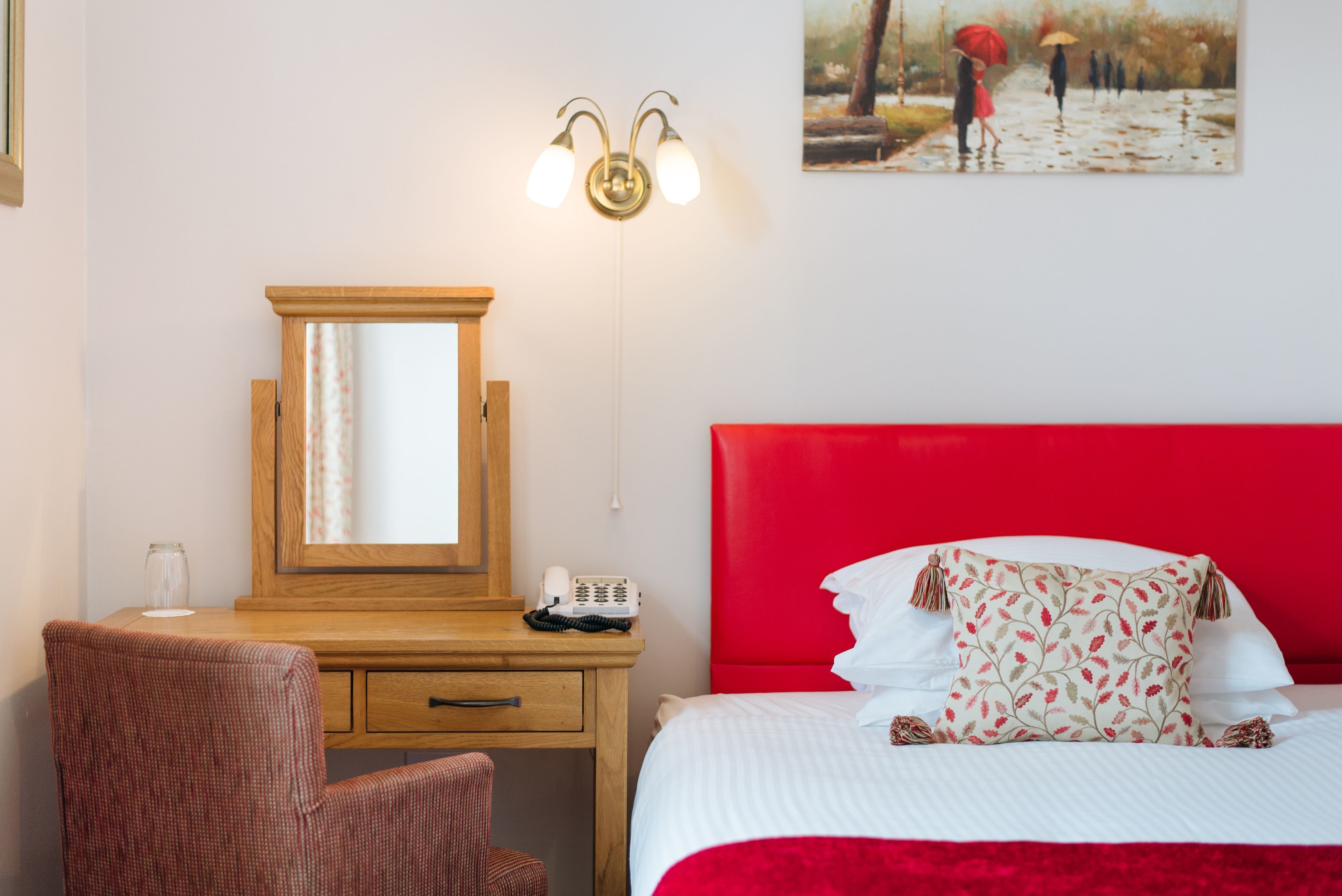 A hotel room with a wooden desk, a red headboard, and a painting of people walking in the rain.