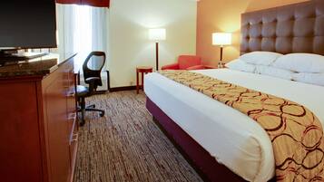 A hotel room with a bed, a desk with a television, a chair, a lamp, and a carpeted floor.