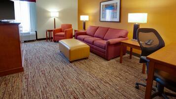 A hotel room with a sofa, ottoman, armchair, desk, and a television.