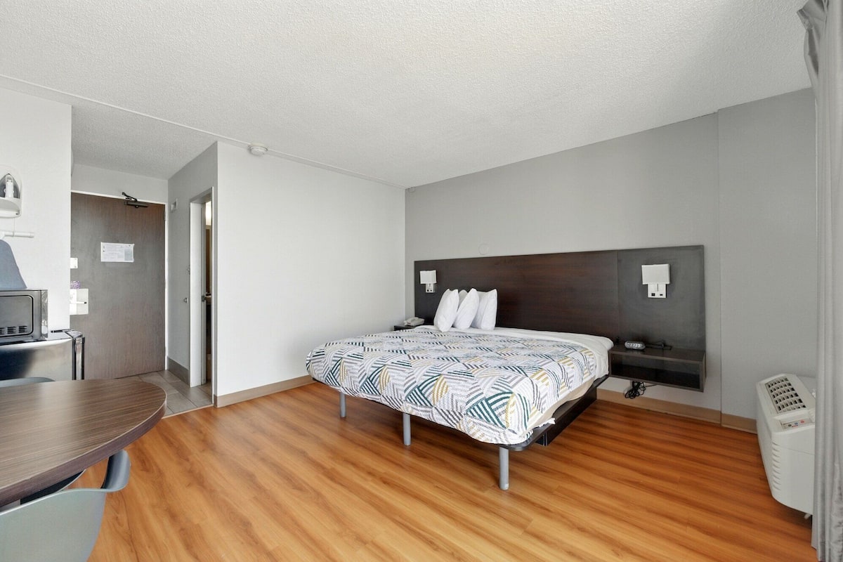 A hotel room with a wooden floor, a bed with a patterned bedspread, a dark headboard, bedside lamps, a desk with a chair, and a heater.