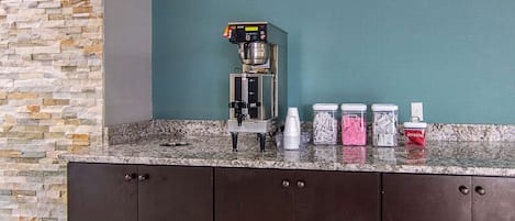 Máquina de café, copos descartáveis e saquinhos de açúcar em uma bancada de granito.