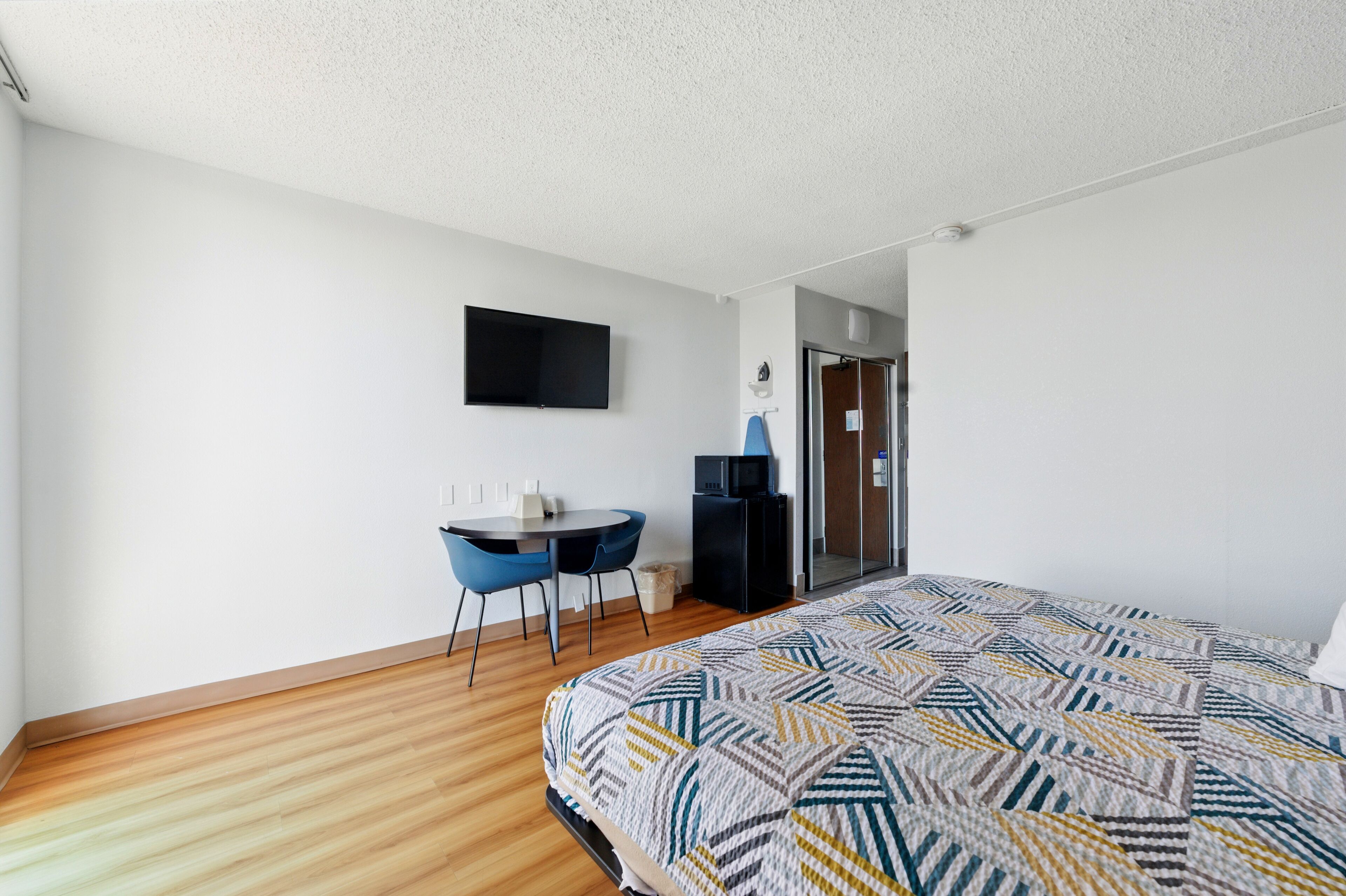 A modern hotel room with a large bed, a desk with two chairs, a flat-screen TV mounted on the wall, and a door leading to a bathroom.