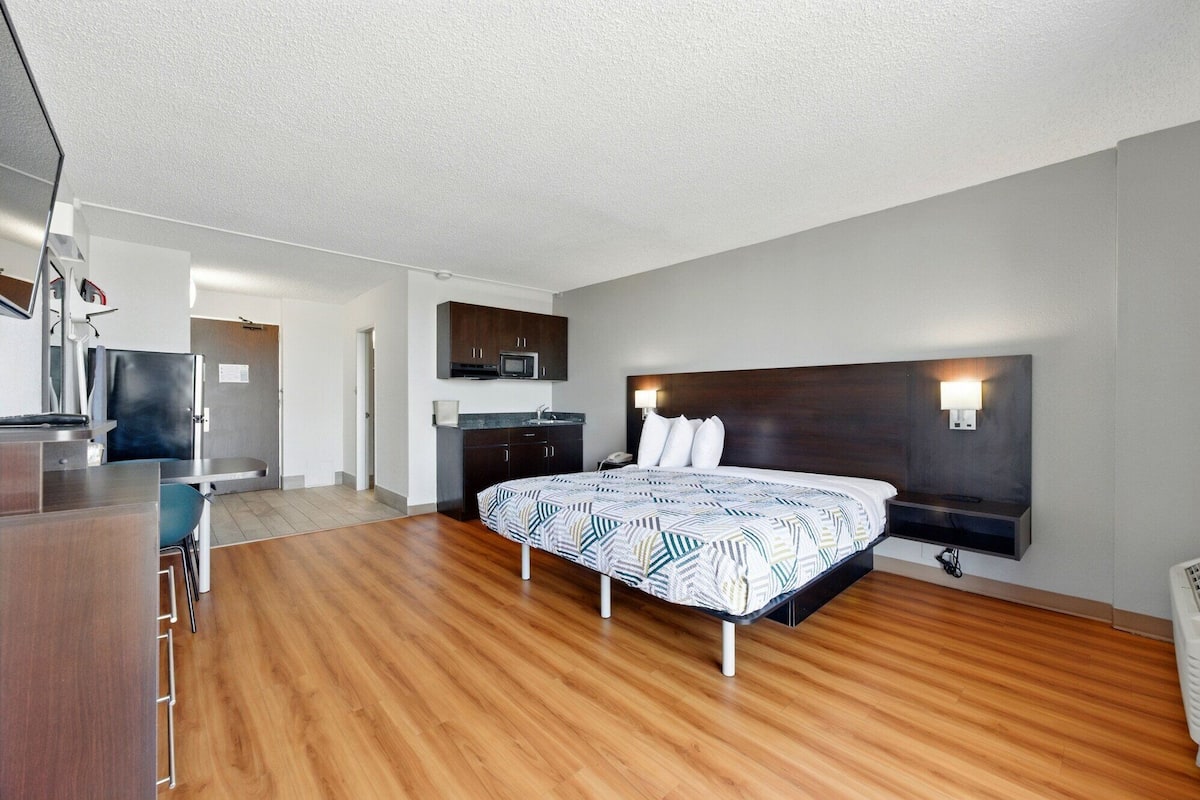 A modern hotel room with a wooden floor, a bed with a patterned bedspread, a dark headboard, a small kitchenette, and a desk with a chair.