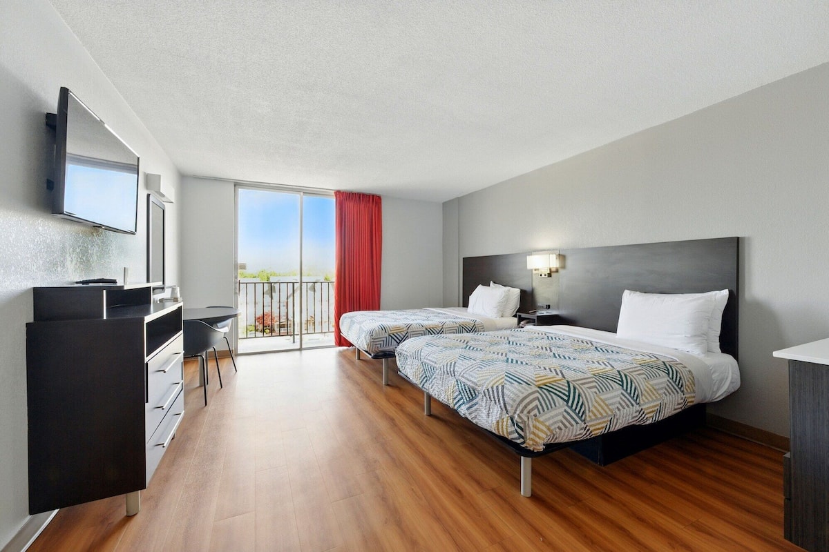 A hotel room with two beds, a desk, a TV, and a balcony with a red curtain.