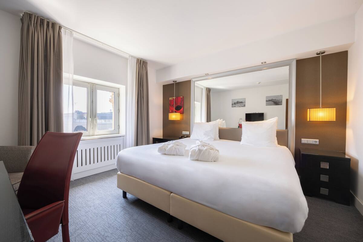 A hotel room with a large bed, a desk, and a chair. There is a window with curtains, a mirror, and a lamp on the nightstand.