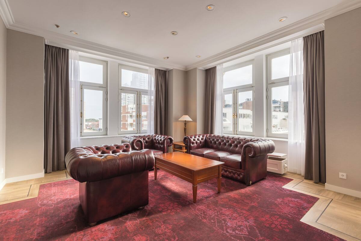 A spacious living room with leather sofas, a wooden coffee table, and large windows offering city views.