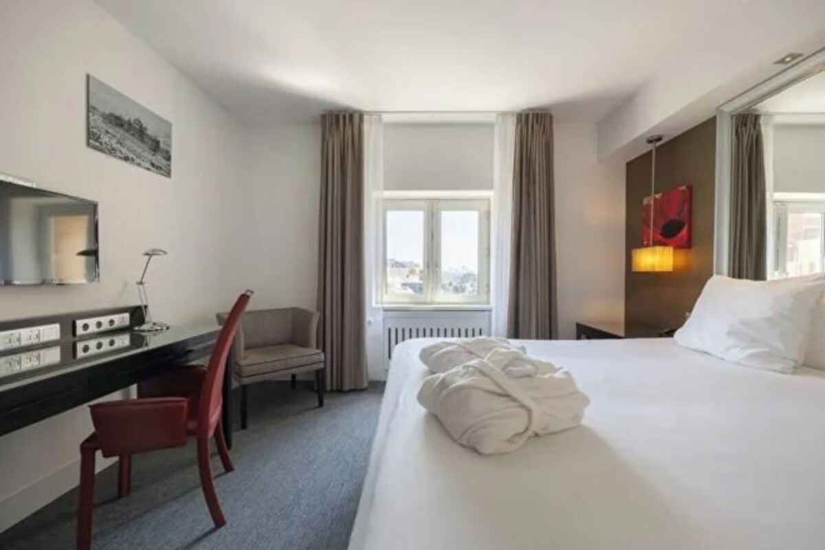 Habitación de hotel con cama, escritorio, silla, ventana y un cuadro en la pared.