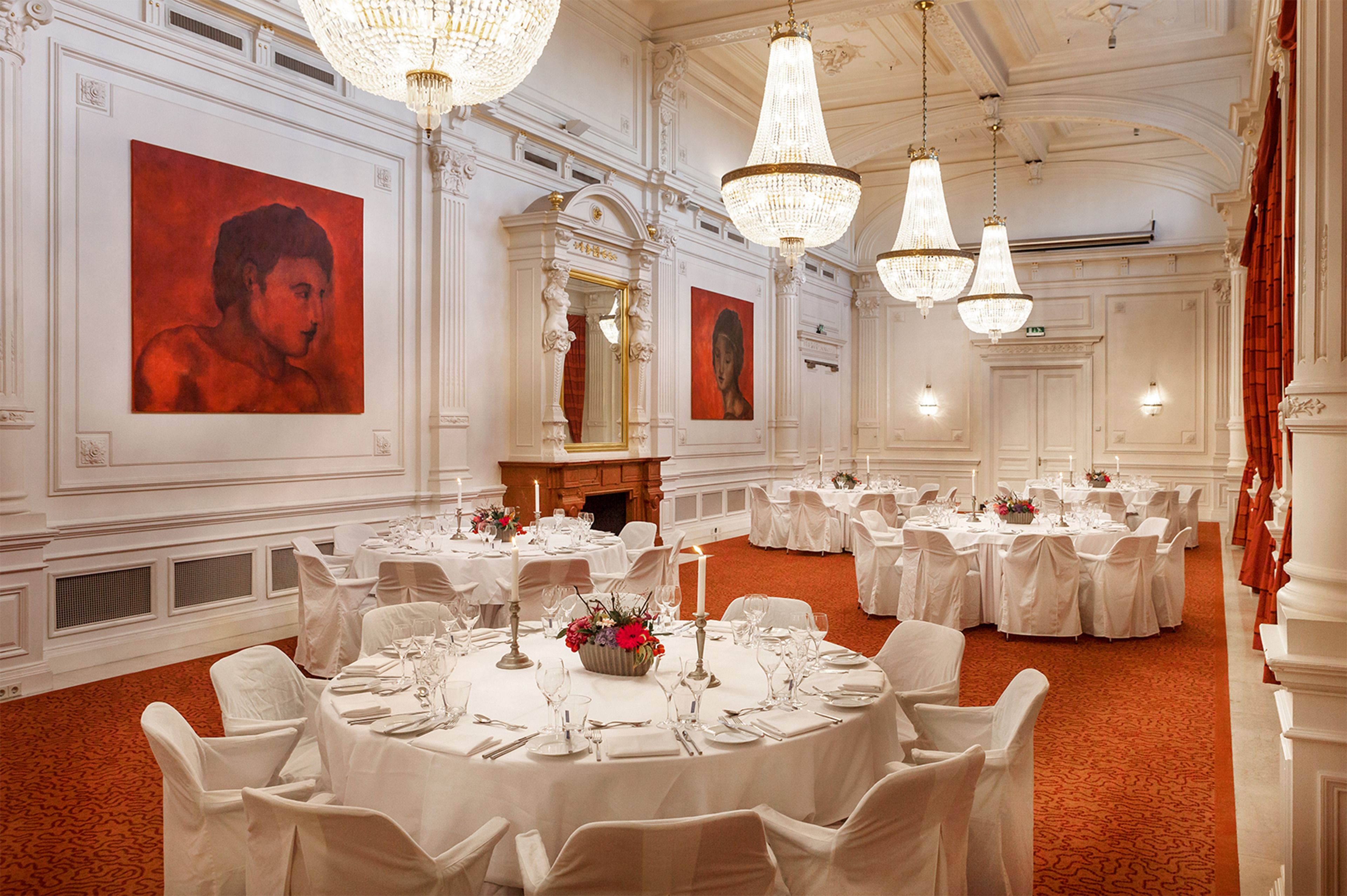 A banquet hall with round tables set for dining, chandeliers, and large framed portraits on the walls.