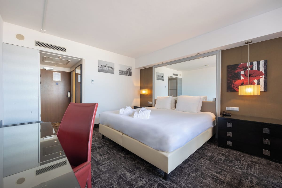 A hotel room with a bed, a desk, a chair, a dresser, a mirror, and artwork on the walls.