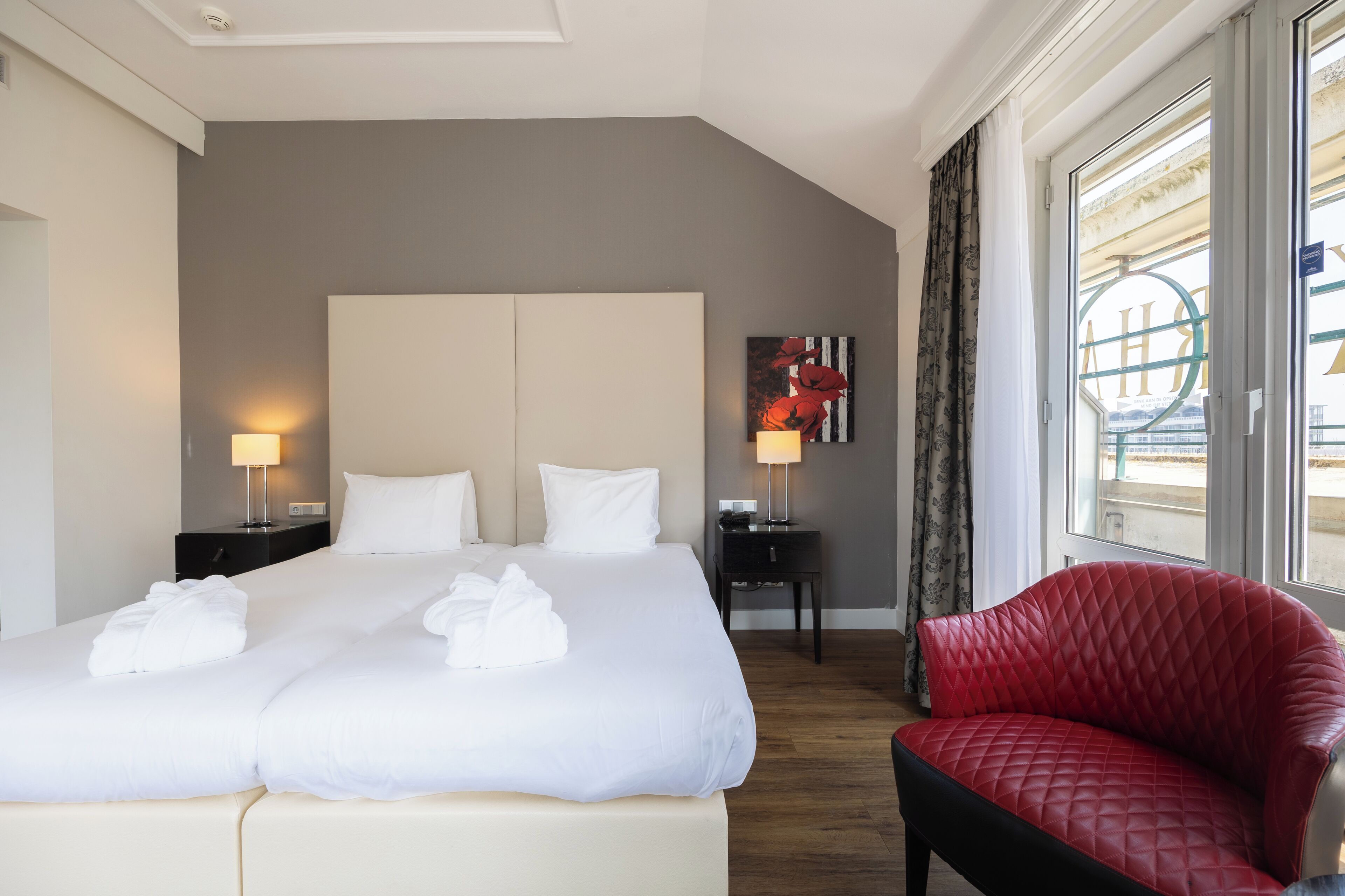A hotel room with a large bed, two bedside tables with lamps, a red chair, and a window with a view of the city.