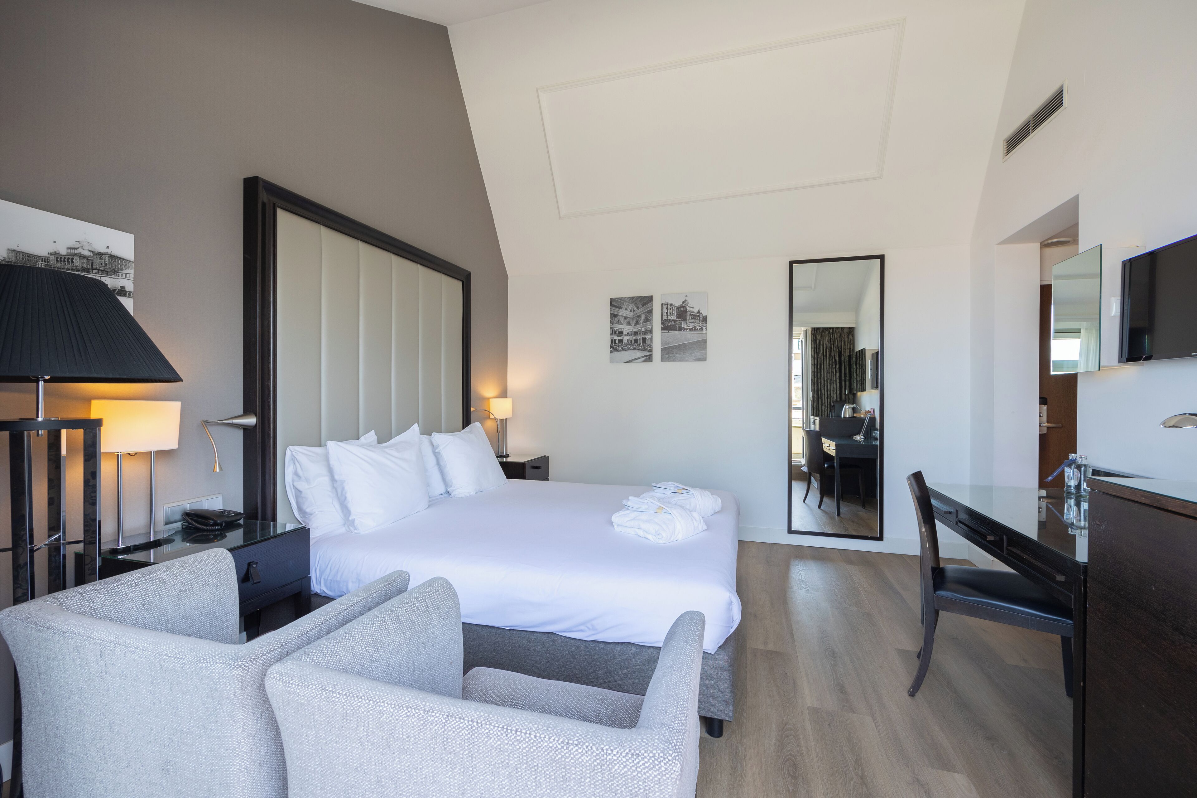 A modern hotel room with a large bed, a desk, a chair, a lamp, and a flat-screen TV.