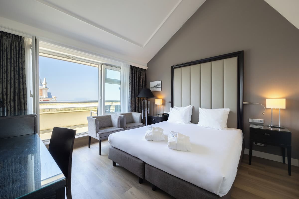 A hotel room with a large bed, a desk, a chair, a nightstand, and a view of a building through the window.