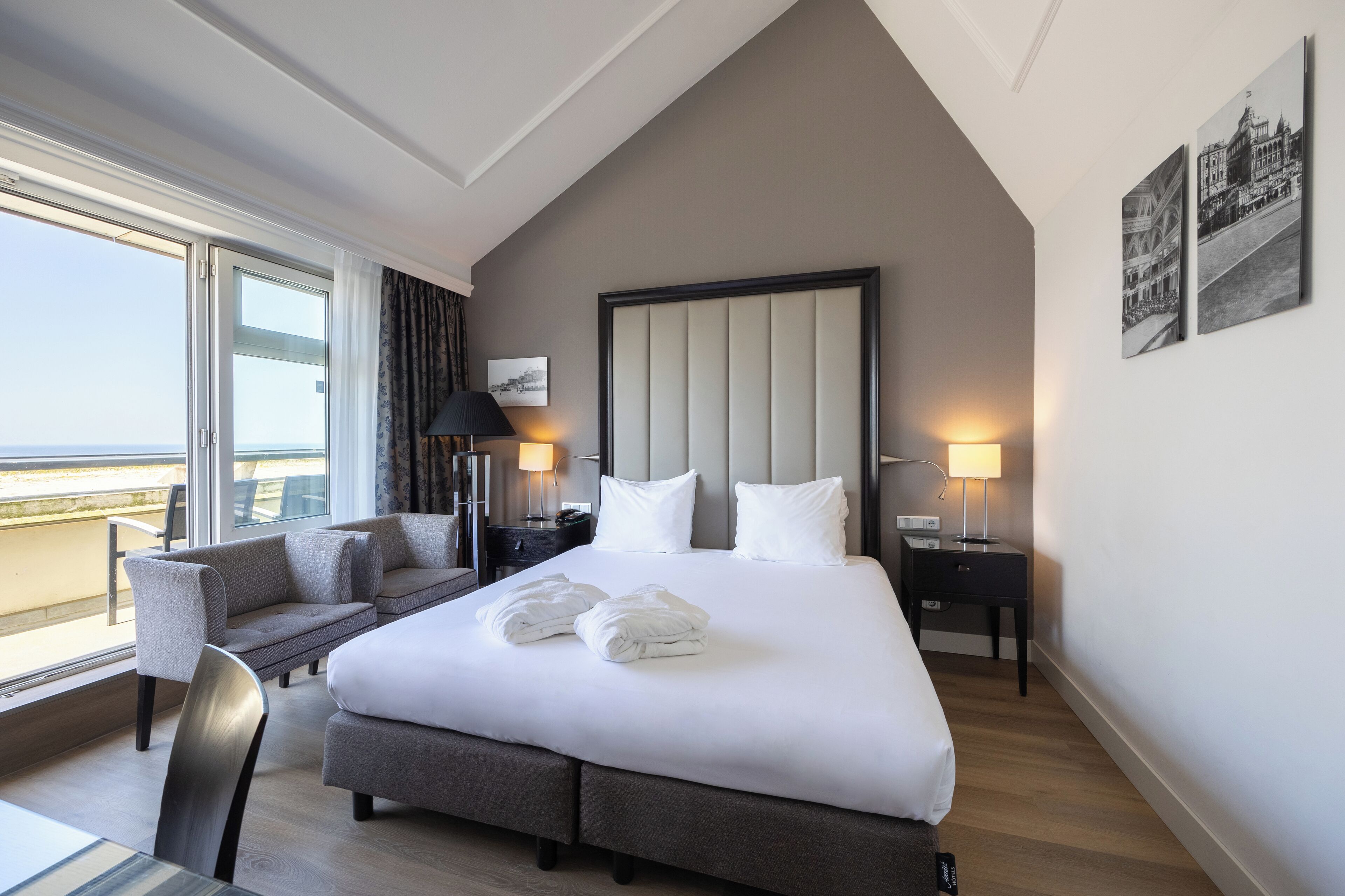 A modern hotel room with a large bed, a sofa, a desk, and a chair. There are two nightstands with lamps. The room has large windows with a view of the sea.