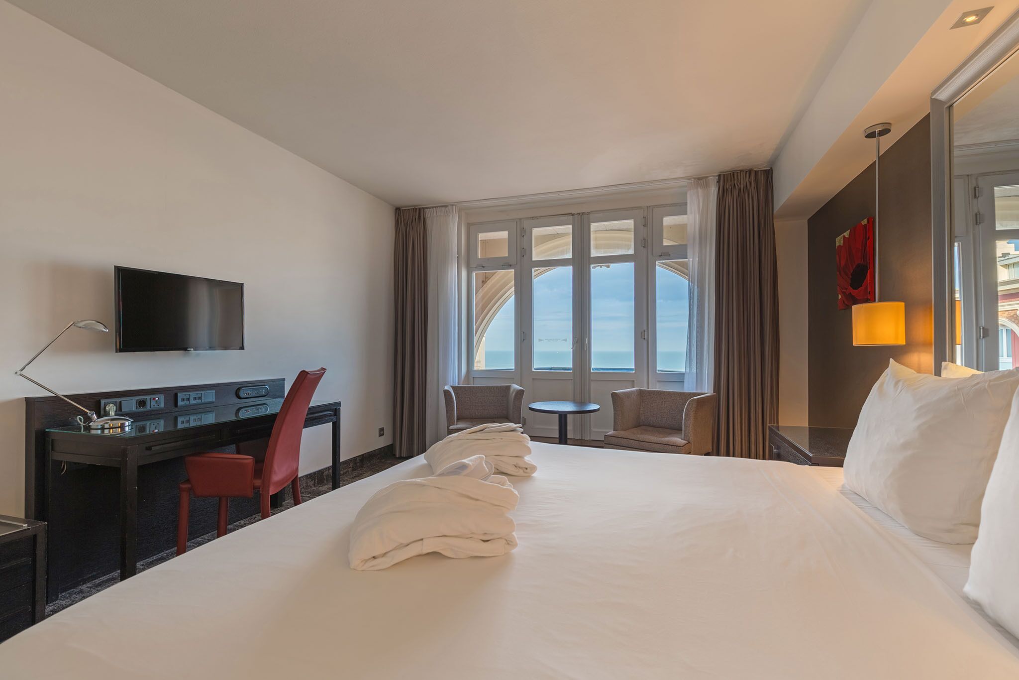 Habitación de hotel con una cama grande, un escritorio con silla, televisor y vista al mar a través de amplios ventanales.