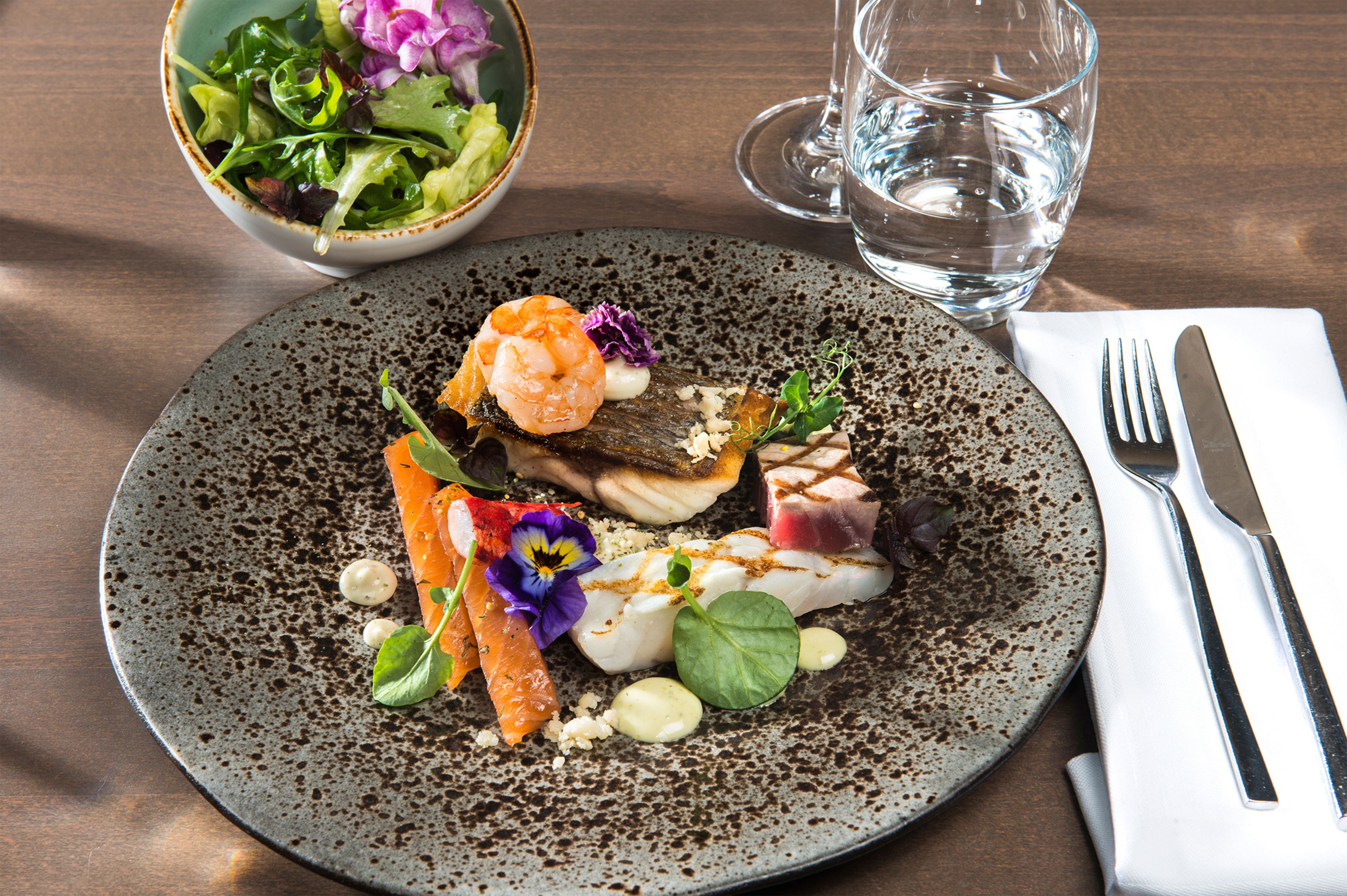 Un plato de comida con diversos platillos, un vaso de agua y un tazón de ensalada sobre una mesa de madera.