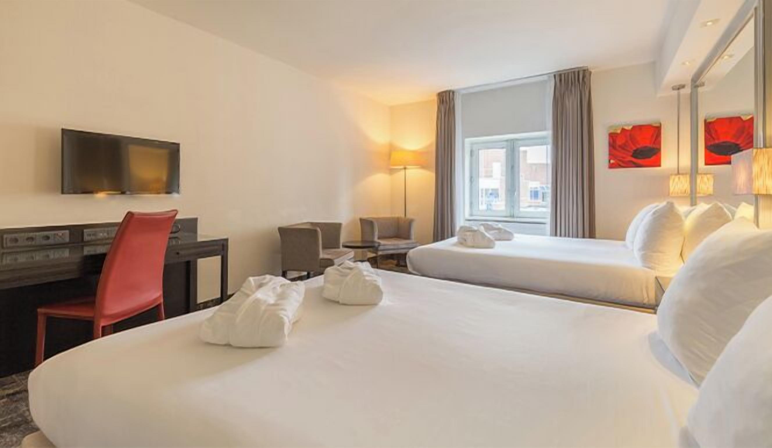 A hotel room with two beds, a desk, a chair, a TV, and a window with curtains.