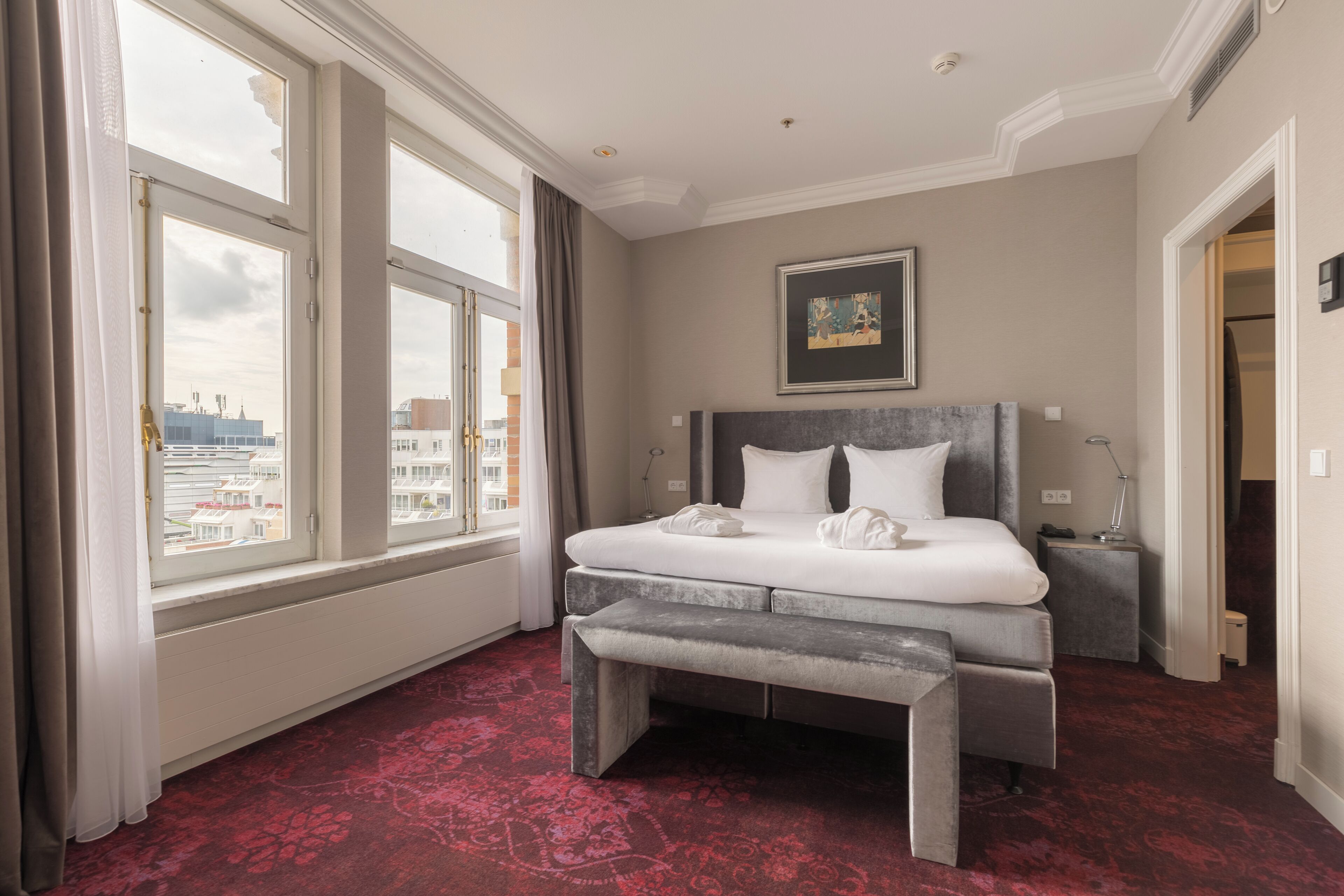 A hotel room with a large bed, a bench, a nightstand, a framed picture on the wall, and a view of the city through large windows.