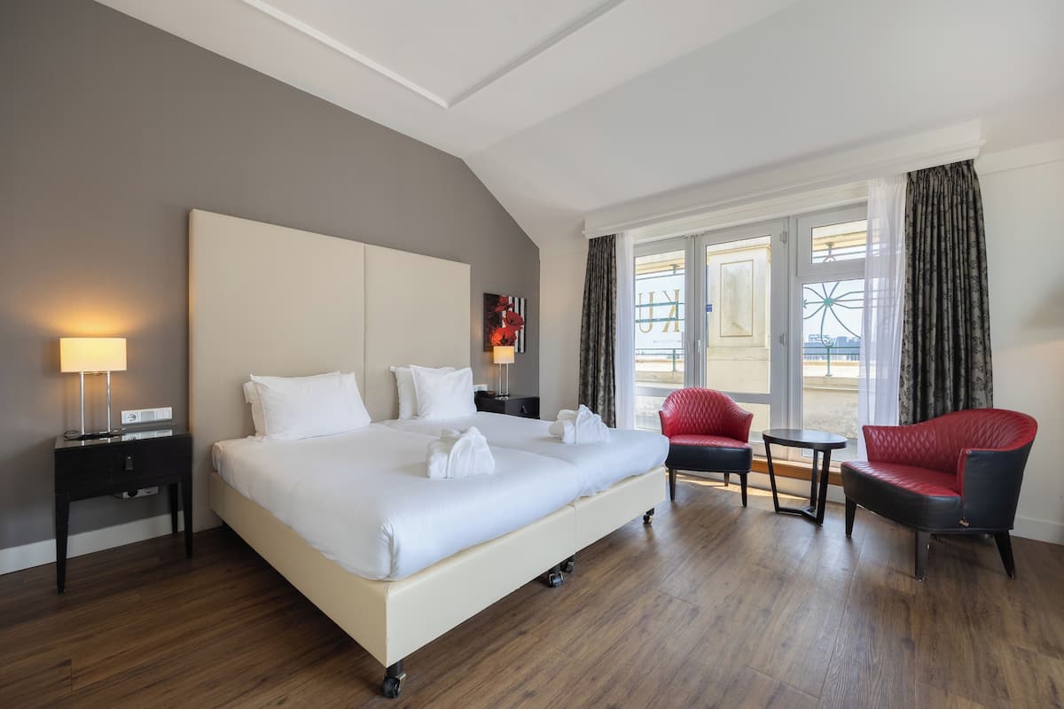 A hotel room with a large bed, a nightstand with a lamp, a red armchair, a small round table, and a view of the outside through a large window.