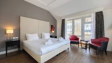Ein Hotelzimmer mit einem großen Bett, einem Nachttisch mit Lampe, einem roten Sessel, einem kleinen runden Tisch und Blick ins Freie durch ein großes Fenster.