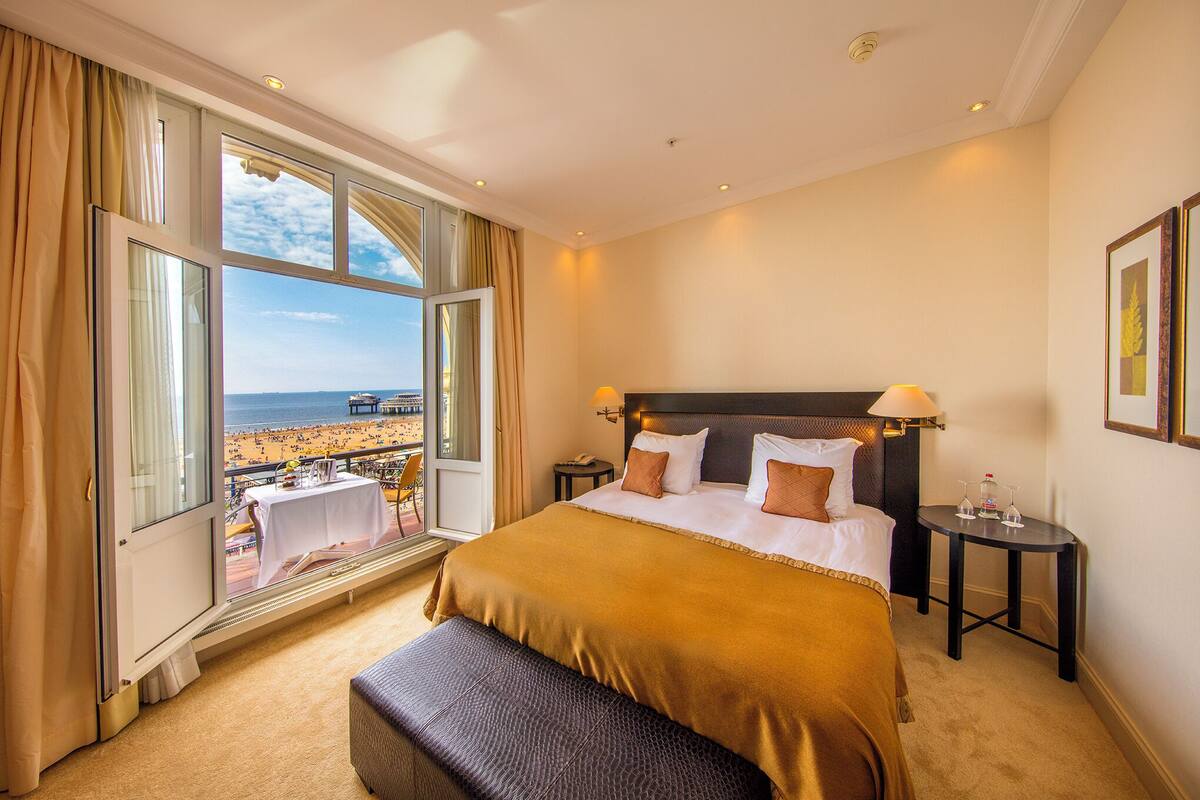 A hotel room with a large bed, a bench, a nightstand, and a view of the beach.
