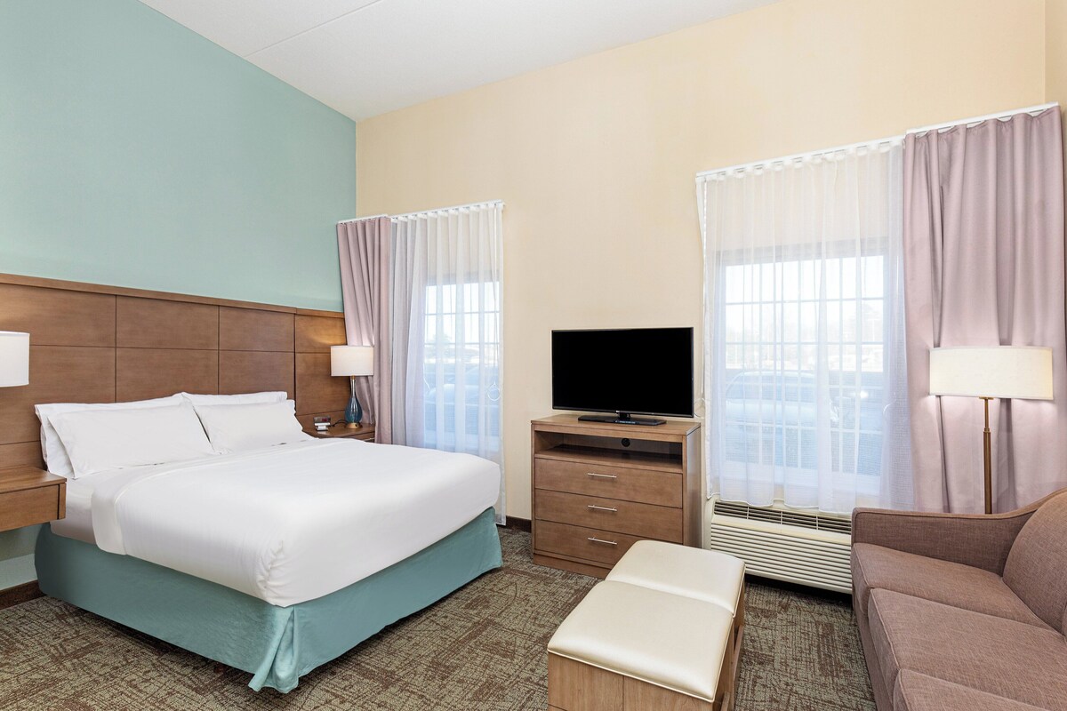 Una habitación de hotel con cama, televisión, escritorio, silla y ventana con cortinas.