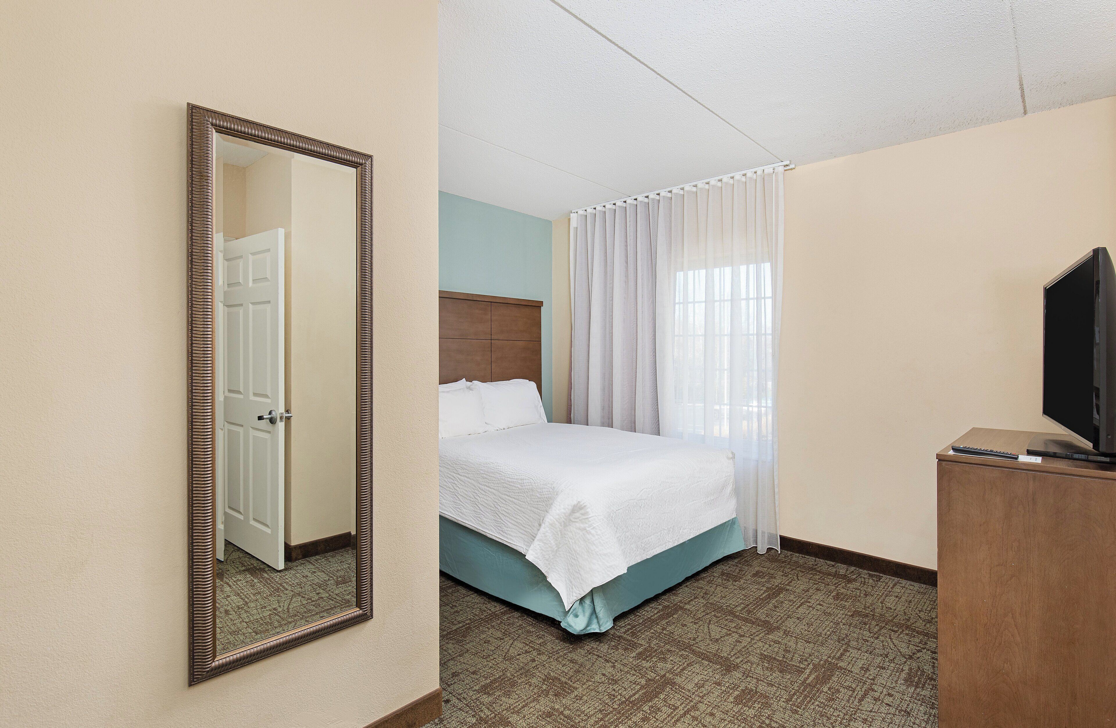 Quarto de hotel com cama, televisão sobre um armário de madeira e espelho na parede.