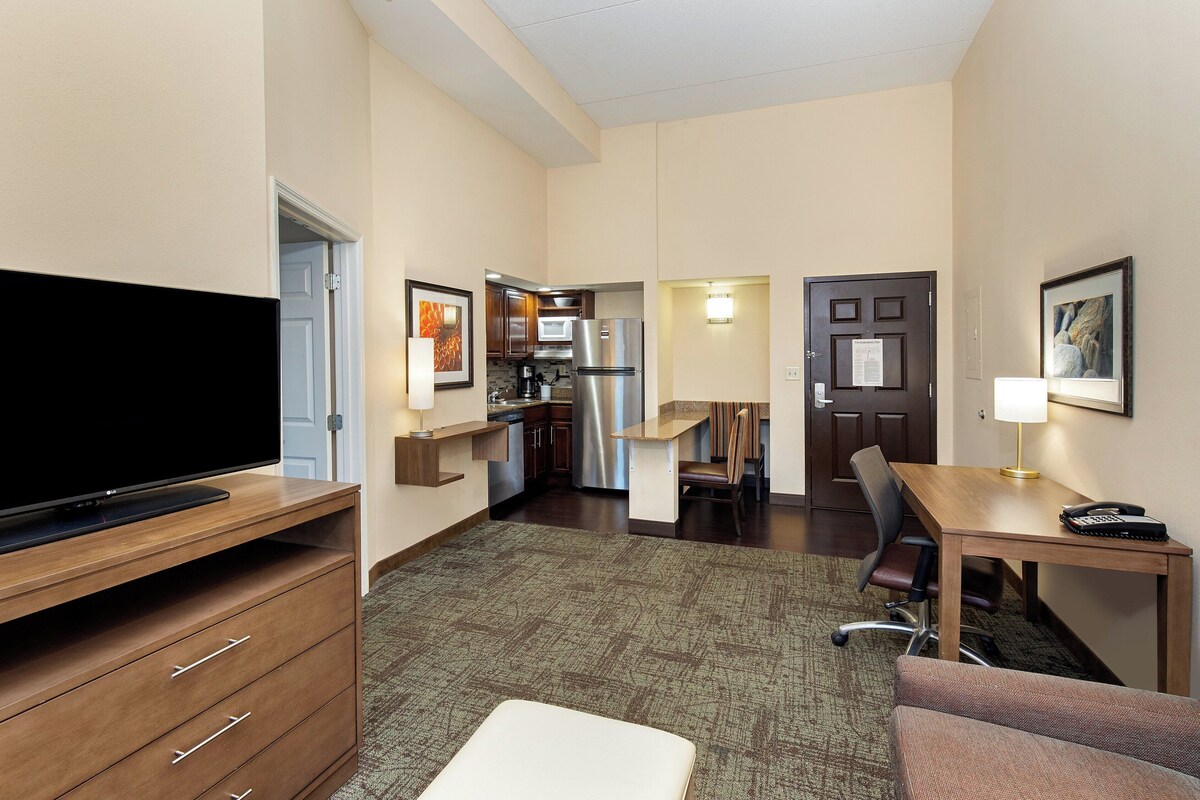 Habitación de hotel con televisión de pantalla plana, un escritorio de madera, una mesa de comedor, una zona de cocina y un área de descanso con sofá.