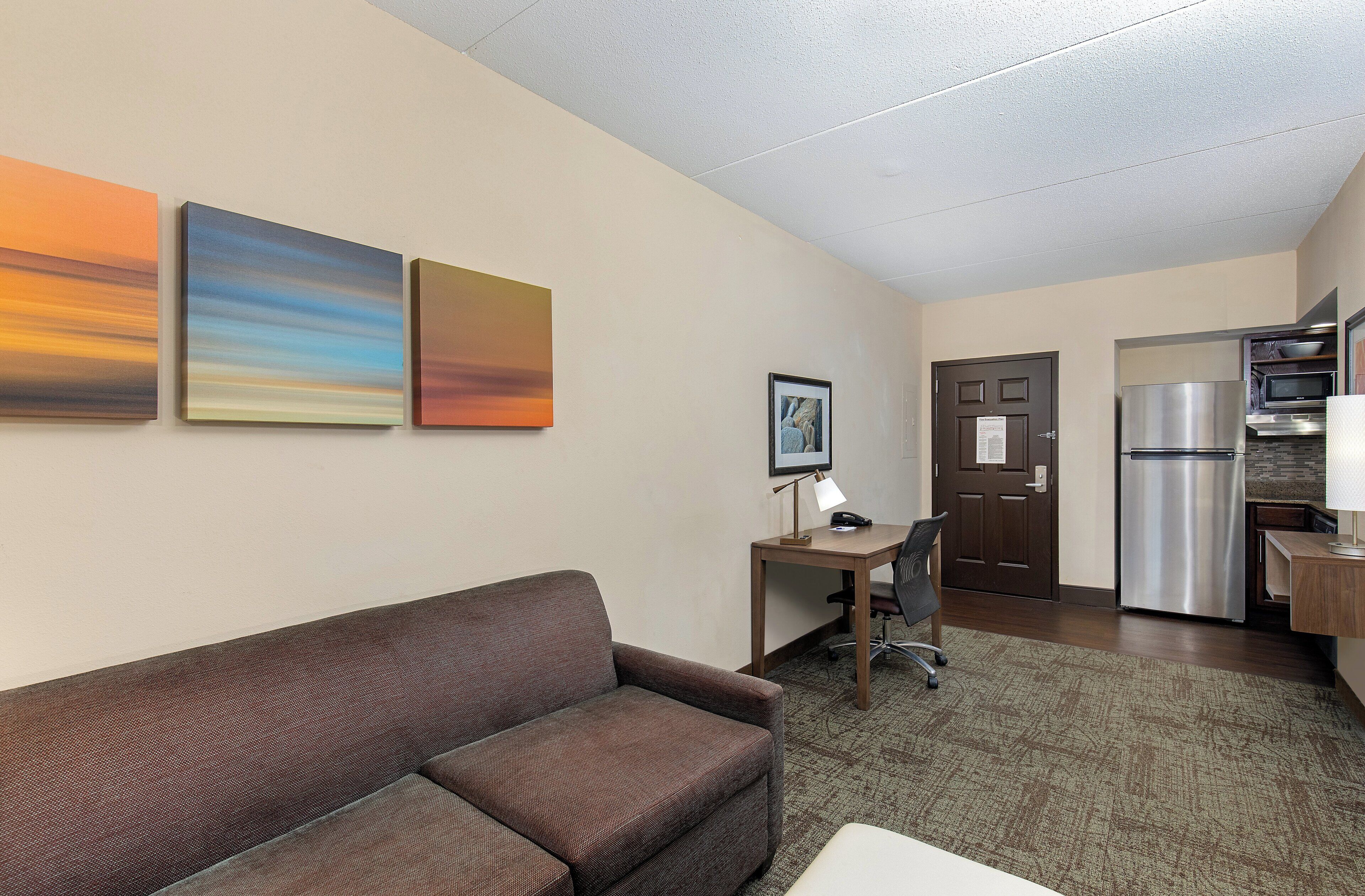 A hotel room with a brown sofa, a desk with a lamp, a framed picture on the wall, and a kitchenette with a refrigerator and microwave.