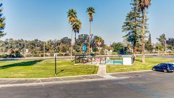 A parking lot with a blue car, a small fenced area with a bench, and tall palm trees.