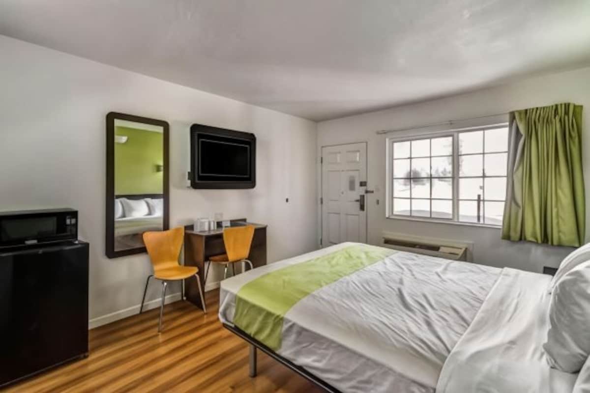 A hotel room with a bed, a desk with two chairs, a television, and a window with curtains.