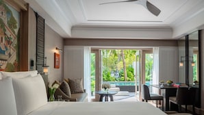 Habitación de hotel con cama, zona de estar con mesa, escritorio y vistas a un jardín y piscina.