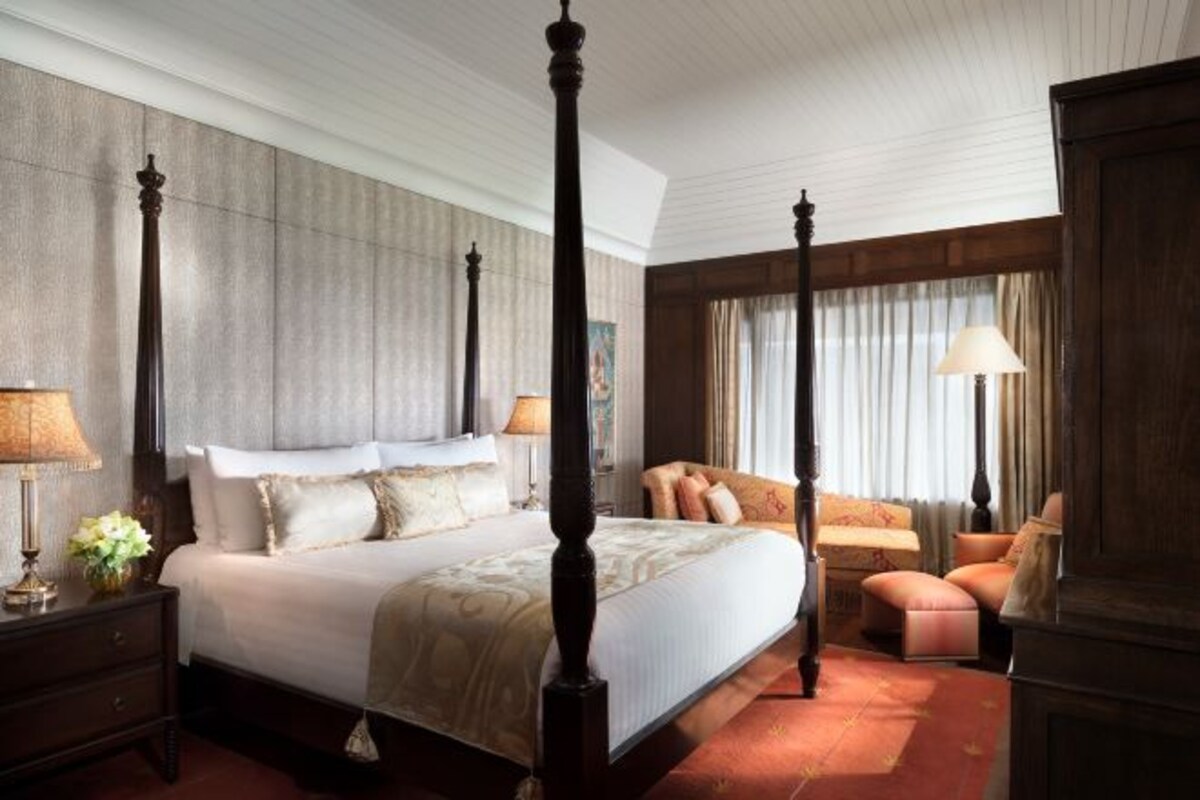 A four-poster bed, a sitting area with a sofa and armchairs, a nightstand with a lamp, and a large window with curtains.