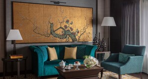 A room with a teal sofa, a matching armchair, a coffee table with a teapot, cups, and a vase of flowers. There is a large painting on the wall and two lamps on either side of the sofa.